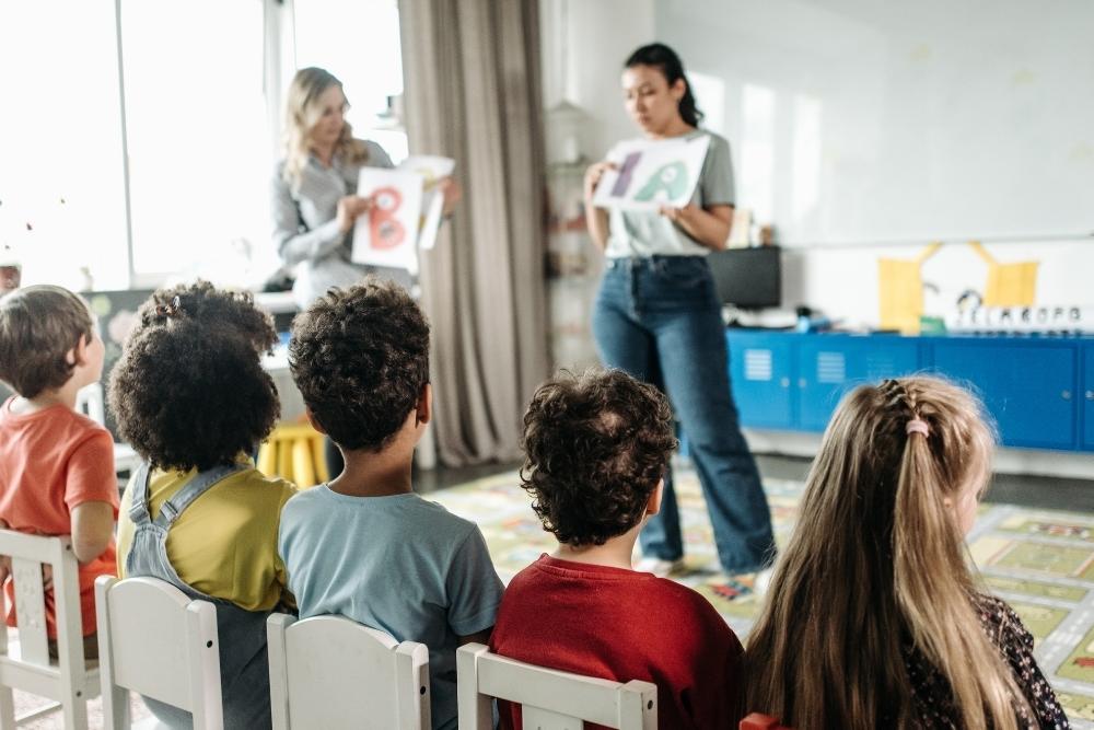 duas professoras mostram letras para crianças sentadas em uma aula de alfabetização.