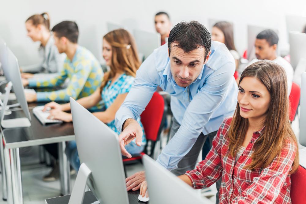 professor auxiliando aluna em sala de informática com outros estudantes ao fundo.