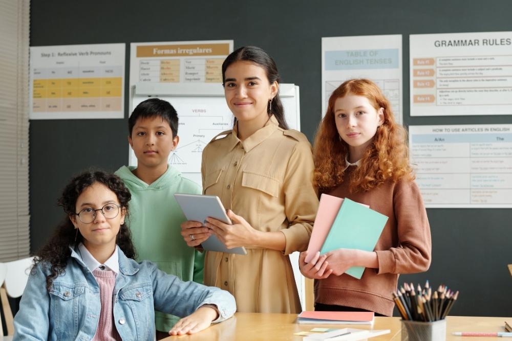 Grupo de alunos e professora em sala de aula participando de atividade do Dia do Estudante, com livros, cadernos e quadro com regras gramaticais ao fundo.