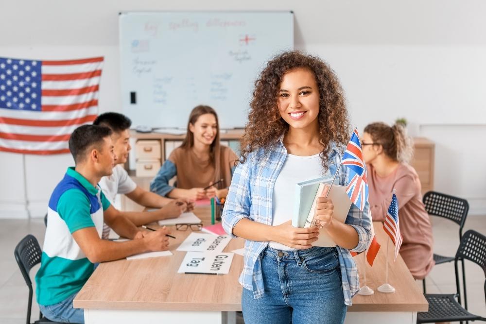 estudante sorridente segura bandeiras do Reino Unido e dos EUA, em sala com colegas e decoração temática de inglês.