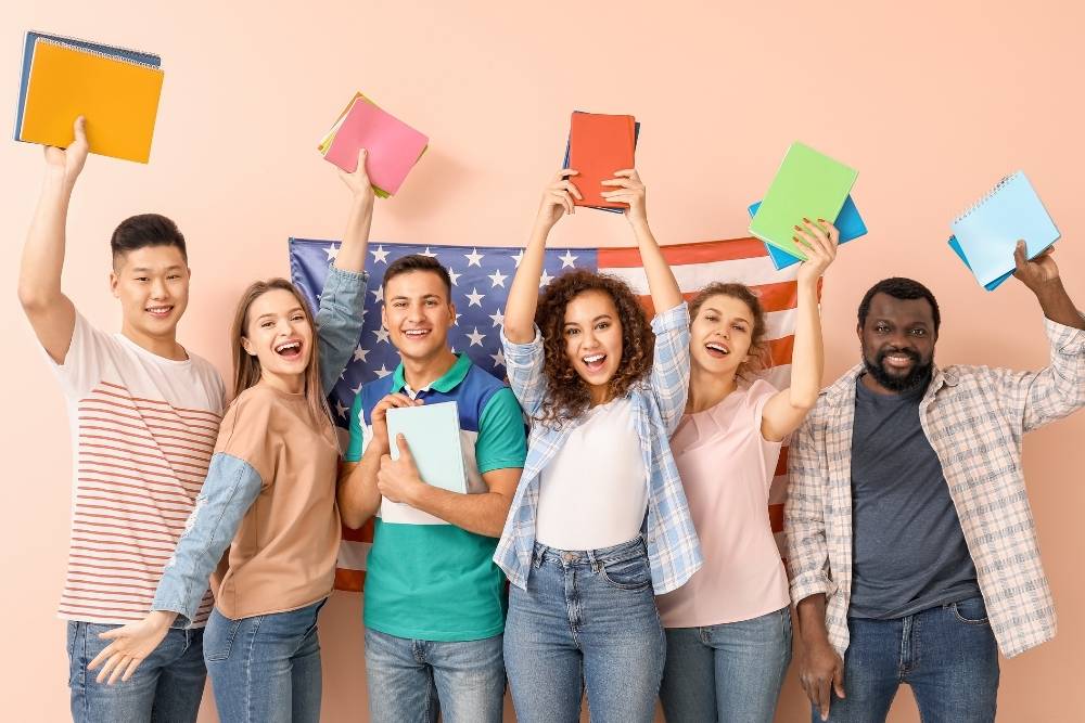 grupo diverso de estudantes felizes levanta cadernos coloridos em frente a uma bandeira dos EUA.