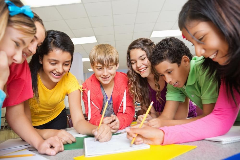 grupo de alunos sorrindo e escrevendo juntos em uma folha sobre a mesa, em ambiente escolar.